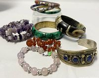 Photo of assorted beaded bracelets and solid bangles showing semi-precious stones like rose quartz, amethyst, lapis lazuli, malachite, and other materials including silver-toned and resin components.