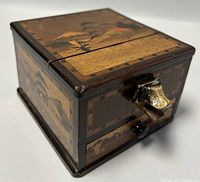 Angle view of the Japanese puzzle box showing the metal animal head handle and landscape inlay.