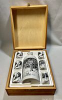 Opened wooden box displaying one ceramic decanter and six matching shot glasses arranged in white foam insert.