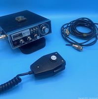 Midland CB radio with microphone and cable on blue background, showing the unit, microphone with push-to-talk button, and cable with connectors.