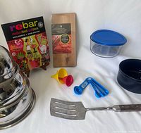 Photo showing full assortment including Rebar cookbook on stand, wooden chopping board, metal spatula, blue measuring spoons, two small funnels, and two plastic storage containers.