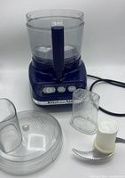 Blue KitchenAid food processor with clear plastic bowl separated next to it, clear plastic lid, clear plastic food pusher, and stainless steel blade attachment.