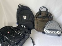 Four backpacks shown together: Swiss Gear black, Adidas black with logo, grey Reebok with large logo, olive Bugatti with handles.