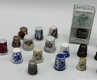 Photo of 15 assorted thimbles arranged on a white background showing porcelain, metal, wooden, and boxed Victoria and Albert 1840 thimble.