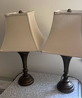 Pair of bronze table lamps on side table with beige square fabric shades, turned off, showing overall condition and style