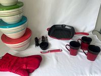 Wide view of the rounded nesting bowls in various sizes with green, blue, and red stripes, red quilted oven mitt, black and red cast iron grill pan, black measuring cups and spoons, red-handled peeler, and three red to black gradient mugs on white background.
