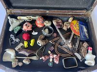 Open wooden case showing assortment of vintage decorative items including figurines, pins, badges, dice, and metal decorative pieces.