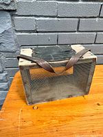 Front and side view of the antique wooden bee box with mesh screen and leather handle on wooden surface in front of brick wall.