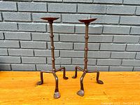 Pair of old wrought iron candlestands standing side by side against a brick wall, showing their overall form and tri-foot base design.