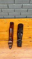 Photo of the front view of two carved African hardwood masks on a wooden table against a gray brick wall.