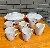 Full view of 2 stoneware casserole dishes with lids and 6 white mugs with black oval design on wooden surface.