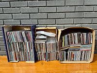 Photo showing three boxes filled with multiple rows of CDs, arranged upright, visible spines with various titles and artists.