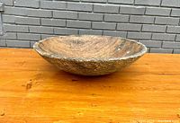 Side angled view of large carved wooden bowl on wooden surface against brick wall background.