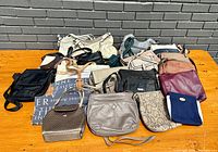 Wide view of fourteen assorted purses and handbags laid out on wooden surface against grey brick wall, showing diverse styles and colors