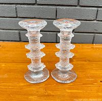 Pair of clear glass Festivo ice candle holders with textured surfaces and ridges, standing on a wooden surface against a brick wall background.