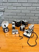 Photo showing full lot of seven vintage cameras arranged on wooden surface against gray brick wall.
