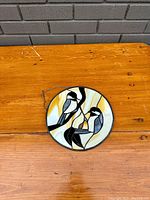 Top-down photo of first stained glass panel on a wooden table showing the detailed birds and branch design with amber and white swirling background and metal chain attached.