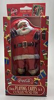 Front view of Coca-Cola Santa-shaped collector tin packaging showing Santa holding Coca-Cola bottles and festive decoration.