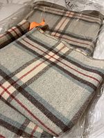 Folded wool blanket showing plaid pattern with cream, brown, light blue, and red colors partially in plastic packaging.