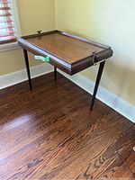 Angled side view of the wooden antique table showing brass handle and three attached wooden legs with one leg missing or detached.