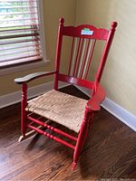 Photo of full red antique vintage rocking chair, showing wooden frame, vertical spindles, armrests, and woven seat with natural material.