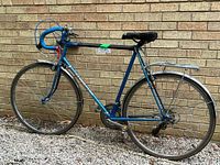 Photo of blue vintage road bike leaning against a brick wall, showing full side view, tires, fenders, and frame details.
