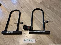 Two black U-lock bike locks lying on wood floor with keys attached and plastic tags, labeled as Lot #17. One lock branded CCM, the other Supercycle with mounting bracket.