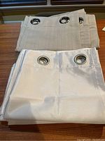 White and beige curtain panels folded on wood table showing metal grommets.