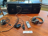 Overview of entire lot showing Sony boom box, black and silver Panasonic Sony headphones, Logitech webcam, Motorola Bluetooth headset, and small MP3 player connected by wires on wooden table.