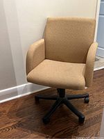 Front view of tan fabric office chair with padded arms and wheeled base on hardwood floor.
