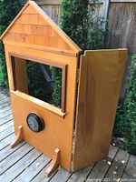 Front angle of wooden puppet theatre showing window stage and clock mounted below. Theatre has house-like roof detail and stands on feet.