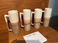 Four handmade pottery mugs arranged side by side on a wooden surface with a note labeled Lot #2 in front, showing their shape, color, and handles.