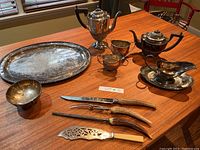 Photo of full tea set on wooden table includes silver serving tray, coffee pot, teapot, creamer, sugar bowl, bone handled carving set, and fish serving utensil.
