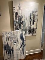 Both abstract cityscape paintings displayed on a wall: one larger vertical canvas featuring a tall skyscraper and city buildings, the smaller canvas leaning against the wall showing complex abstract architectural elements.