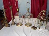 Full view of the two glass and brass lamps, two gold-colored candle holders, and brass and glass chandelier on white surface with red curtain background.
