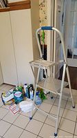 Metal step stool with blue accents alongside various cleaning supplies placed on tiled floor near white cabinet.