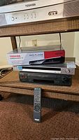 All three electronic devices shown on a shelf. Toshiba SD3300 DVD player in sealed box atop Sony DVD/CD player and Sharp VCR player, with remote placed in front.