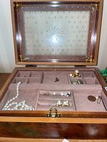 Open burled wood veneer jewelry box with compartments holding pearl necklace and several pearl earrings and pins; glass top with decorative etching visible