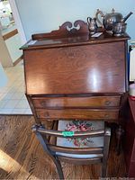Full view of wood secretary desk with matching chair in front. Desk has drop-front surface, two drawers, and decorative carved scroll top. Chair has floral cushioned seat.