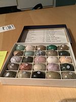 Top-down and angled views of a compartmentalized box containing 25 polished stone specimens, each numbered with a small white circular sticker.