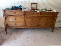 Full front view of vintage wood dresser showing all nine drawers and tapered legs on carpeted floor, top surface holding various personal items.