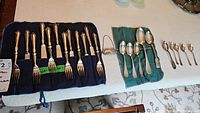 12-piece Walker and Hall flatware set with forks and cutters displayed in a blue protective roll, alongside 5-piece RYRIE spoon set, 4 unmarked sugar spoons, and a Scotch bar bottle tag.