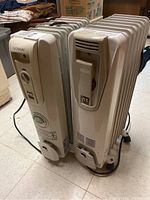 Pair of oil-filled radiator heaters side by side on a linoleum floor, showing entire heaters and power cords.