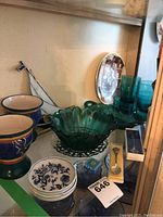 Displayed teal glass bowls with scalloped edges and sunflower pattern, blue glass cups, pottery goblets, and stained glass dual boat decoration in a shelf.