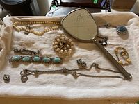 Overview photo of assorted vintage costume jewelry pieces spread on cloth including necklaces, bracelets, rings, and a vintage hand mirror.