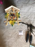 Full view of the wooden cuckoo clock mounted on the floor, showing two pinecone-shaped weights connected by tangled chains and the colorful clock face with painted leaf decorations.