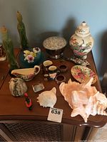 Wide angle photo showing a table with an assortment of decorative items including seashells, porcelain flowers, Asian-inspired candle figures, colored glass plate, Royal Doulton heads, floral plates, and small figurines.