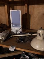 White Vertux light therapy lamp, beige desk lamp with conical shade, small lens light on wooden shelf under dim lighting.