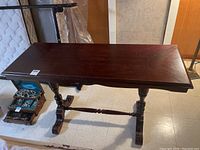 Image showing the dark wood rectangular hallway table with carved legs and a cross beam support, placed on a light tile floor with some items visible behind and below.