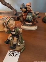 Photo of both Hummel figurines on a wooden surface showing one seated boy and one two children figurine, with the hand number card 637.